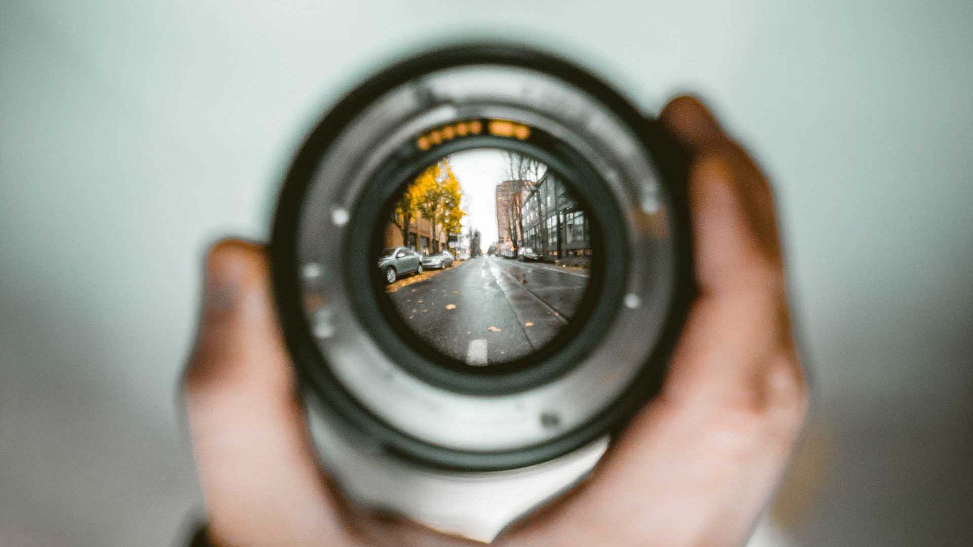 man holding camera lens. the background is blurry. a city street is in focus through the lens.