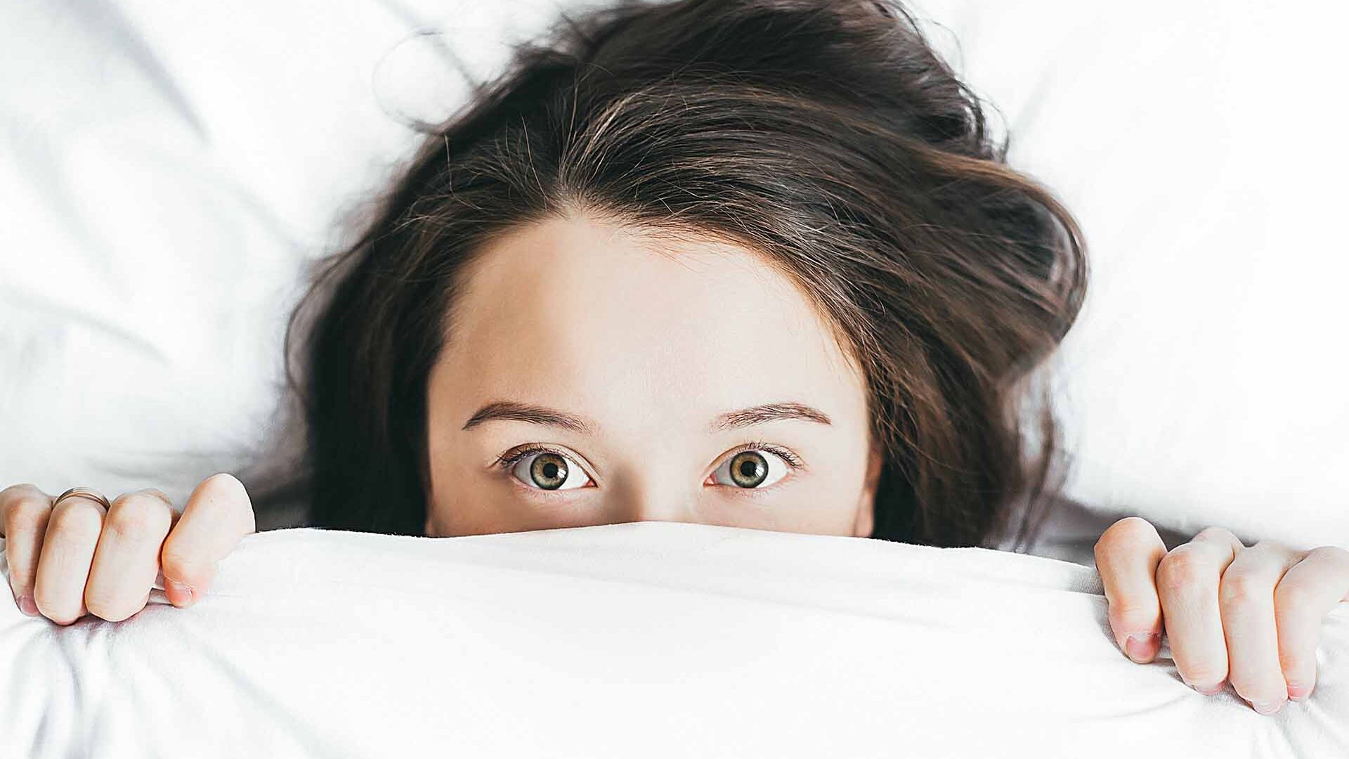 woman in bed with the covers over part of her face. she appears to be hiding.