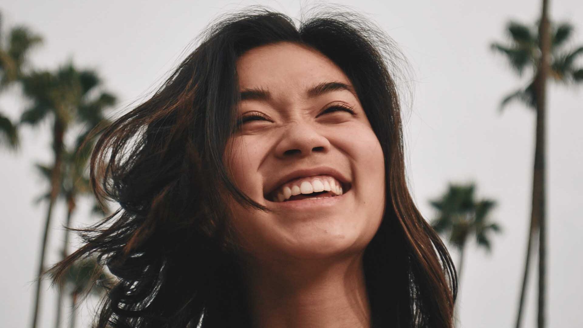 young Asian woman with big smile on her face. she is standing in the street with palm trees behind her.